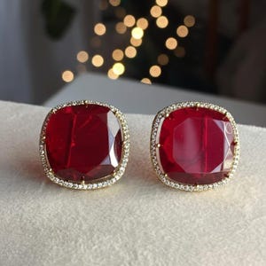 Peut inclure: Deux boucles d'oreilles carrées en pierres précieuses rouge foncé. Chaque boucle d'oreille est entourée d'un halo de petites pierres claires scintillantes serties dans un métal doré. Les boucles d'oreilles sont présentées sur une surface claire.