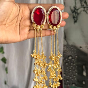 May include: A pair of gold-colored earrings featuring large, oval, deep red gemstones surrounded by small white pearls. Long, dangling chains with clusters of small gold beads hang from the earrings, creating a flowing, elegant design.