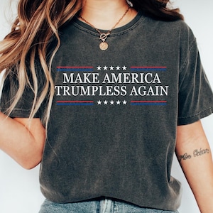 Puede incluir: Camiseta gris oscuro con el texto "MAKE AMERICA TRUMPLESS AGAIN" en blanco, con detalles en rojo y azul. La camiseta se combina con vaqueros azules y zapatillas blancas. La imagen incluye el logotipo de TeeCraftStoreCo.