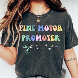 May include: Dark gray t-shirt with the words "FINE MOTOR PROMOTER" in colorful, retro-style lettering. The shirt features a pair of scissors and star graphics. The shirt is worn with blue jeans and white shoes.