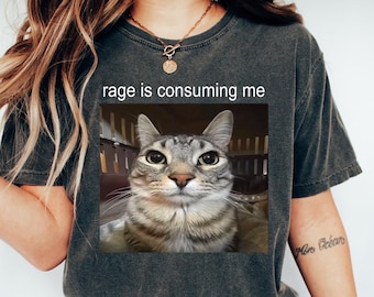 May include: Dark gray t-shirt with the text "rage is consuming me" above a cat image. The cat has gray and brown stripes. The shirt is paired with blue jeans and white sneakers.