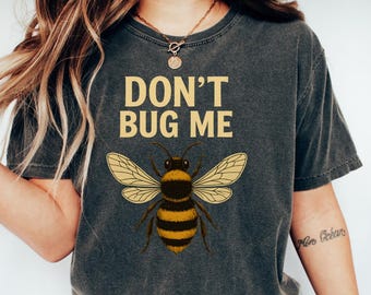 May include: Dark grey t-shirt featuring the text "DON'T BUG ME" and a detailed bee illustration. The bee has yellow and black stripes and wings. The shirt is worn with blue jeans and white trainers.
