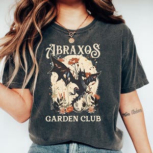 Puede incluir: Camiseta gris oscuro con el texto "ABRAXOS GARDEN CLUB". El gráfico presenta un dragón negro con elementos florales. La camiseta se usa con vaqueros azules y zapatillas blancas.