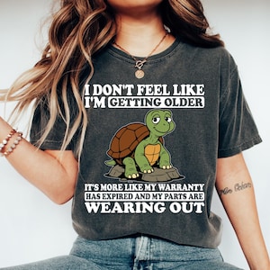 May include: Dark gray t-shirt with the text "I DON'T FEEL LIKE I'M GETTING OLDER" and "IT'S MORE LIKE MY WARRANTY HAS EXPIRED AND MY PARTS ARE WEARING OUT". The shirt features a cartoon turtle graphic. The brand name "TeeCraftStoreCo" is visible.