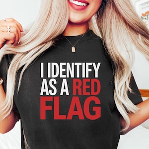 May include: Black crew neck t-shirt featuring the text "I IDENTIFY AS A RED FLAG" in white and red. The shirt is a casual style.