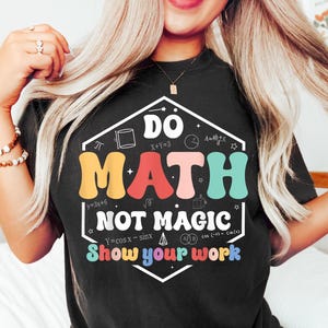 May include: Black t-shirt with the words "DO MATH NOT MAGIC" in a colorful, retro font. The shirt also includes mathematical symbols and the phrase "Show your work".