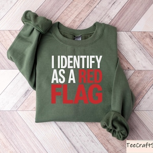 May include: An olive green sweatshirt with the text "I IDENTIFY AS A RED FLAG" in white and red lettering. The sweatshirt is folded on a light brown and white wooden surface.