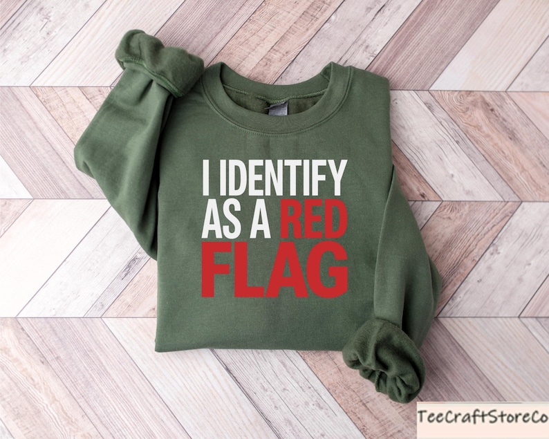 May include: An olive green sweatshirt with the text "I IDENTIFY AS A RED FLAG" in white and red lettering. The sweatshirt is folded on a light brown and white wooden surface.