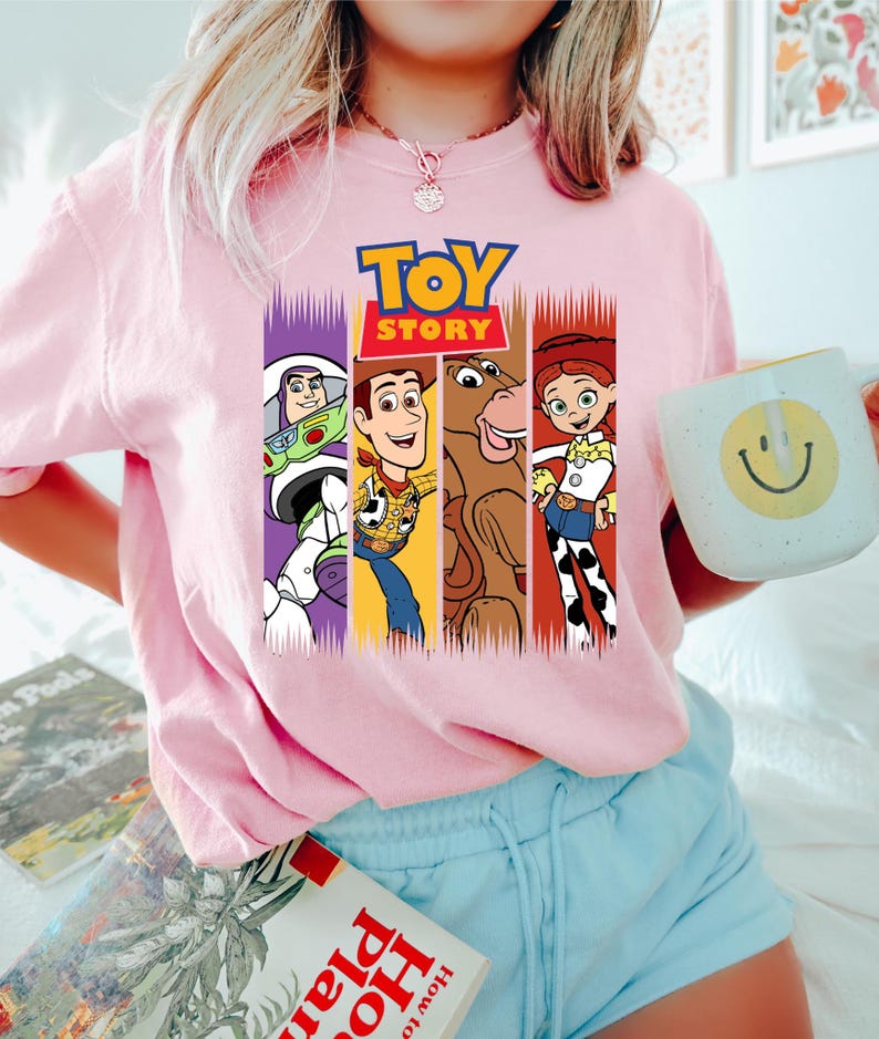 以下が含まれることがあります： ピンク色のクルーネックTシャツ。カラフルなトイ・ストーリーのキャラクターと映画のロゴがプリントされています。デザインには、バズ・ライトイヤー、ウッディ、ブルズアイ、ジェシーが含まれています。黄色のスマイリーフェイスの白いマグカップが手に持たれています。