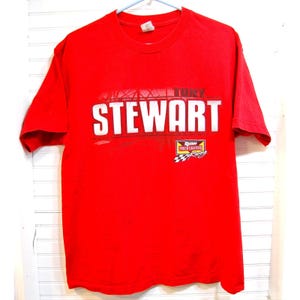 May include: A red t-shirt featuring the name "STEWART" in large white letters. Above the name is the name "TONY" and below is the "RUSH TRUCK CENTERS" logo with a checkered flag. The shirt is hanging on a white wall.