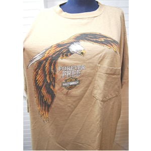 May include: Tan t-shirt featuring a large graphic of an eagle with outstretched wings. The eagle is depicted in shades of brown, black, and white. The text "FOREVER FREE" and "HARLEY-DAVIDSON CYCLES" are printed below the eagle. A pocket is on the left side.