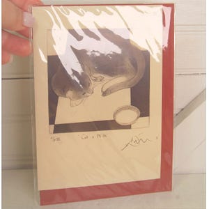 May include: A greeting card featuring a sepia-toned illustration of a cat curled up near a bowl. The artwork is titled "Cat o' Milk" and includes an artist's signature. The card is encased in a clear plastic sleeve with a red border.
