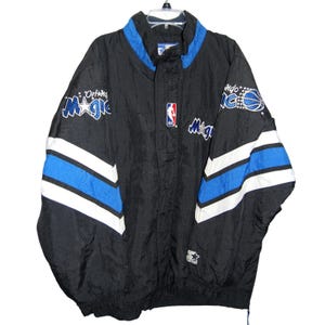 Op de afbeelding: Zwarte Orlando Magic NBA-jas met blauwe en witte accenten. Voorzien van het teamlogo op de borst en mouwen, en het NBA-logo. De jas heeft een Starter-logo aan de onderkant.