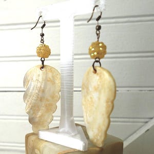 May include: A pair of drop earrings featuring large, leaf-shaped, iridescent cream and gold shell pendants. Each earring is accented with a single, textured, round yellow bead positioned above the shell, suspended from a dark metal hook.
