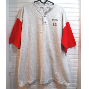 Puede incluir: Una camiseta Henley gris con mangas cortas rojas. La camisa tiene tres botones y un pequeño gráfico de dos banderas a cuadros y el número 12 en rojo. La camisa está colgada de una percha negra.