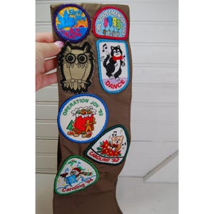 Pode incluir: Uma faixa marrom de escoteira com vários patches bordados. Os patches incluem uma coruja, um husky dançando e desenhos temáticos de feriados. A faixa é exibida em um fundo branco.