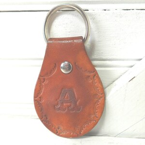 May include: A teardrop-shaped brown leather keychain featuring a stamped letter A in the center. The leather has a decorative embossed border pattern and is secured with a silver-toned metal rivet. A circular silver-toned metal split ring is attached.
