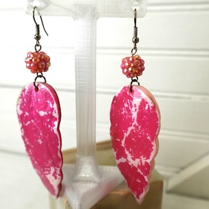 Pode incluir: Um par de brincos pendentes com grandes pingentes em forma de folha com um padrão marmorizado vibrante em rosa choque e branco. Cada brinco é rematado com uma pequena conta redonda facetada vermelho-alaranjada e um gancho de metal escuro.