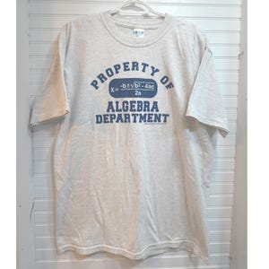 May include: A light gray short-sleeve t-shirt featuring blue text that reads Property of Algebra Department with the quadratic formula printed in a blue oval. The shirt has a crew neckline and is displayed on a white plastic hanger.