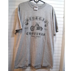 May include: A grey t-shirt with a motorcycle graphic and the text "Sturgis Choppers South Dakota." The design includes a banner with the word "Sturgis" above the motorcycle. The shirt is hanging on a black hanger against a white-painted wall.