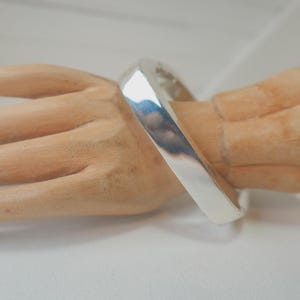 May include: A polished silver bangle bracelet with a smooth, reflective surface and a wide, flat band design. The bracelet features a clean, minimalist aesthetic with a continuous circular shape, showcasing a bright metallic finish that catches light.