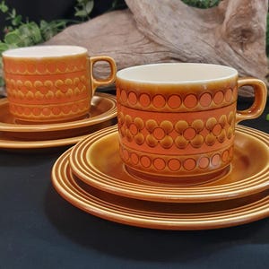 May include: Two sets of vintage orange ceramic teacups and saucers. The cups and saucers have a floral and circular pattern. The cups have a handle and are sitting on top of the saucers. The set is on a black surface.