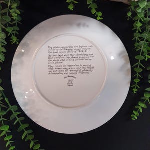 May include: A white decorative plate with a faded, cloudy border. The plate features handwritten text commemorating the role of the Bingley miners' wives during the 1984-5 strike. The text highlights their hard work, cheerfulness, and sacrifice.