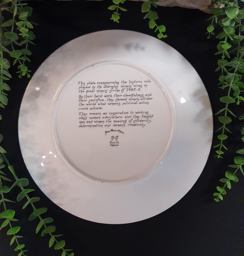 May include: A white decorative plate with a faded, cloudy border. The plate features handwritten text commemorating the role of the Bingley miners' wives during the 1984-5 strike. The text highlights their hard work, cheerfulness, and sacrifice.