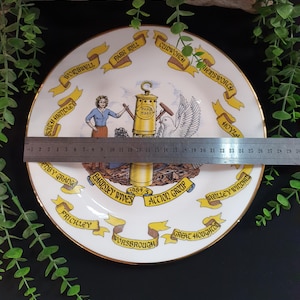 May include: A decorative white plate with a gold rim, featuring a central illustration and surrounding banners with text. The plate has a diameter of approximately 10 inches. The illustration includes a woman, a dove, and a lamp post.
