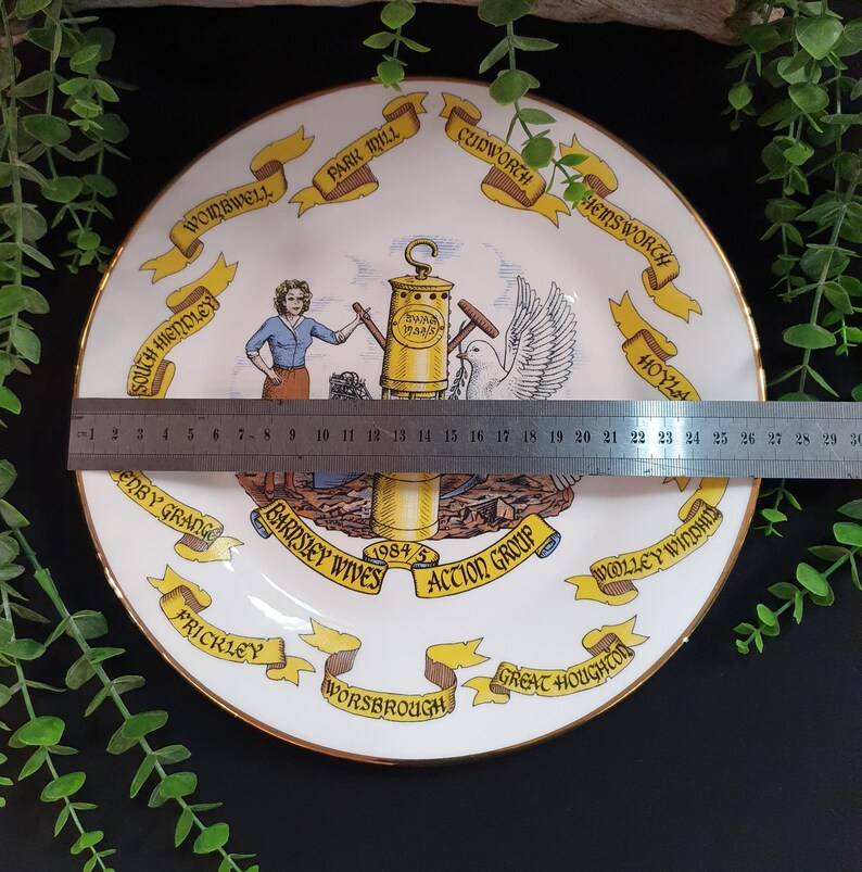 May include: A decorative white plate with a gold rim, featuring a central illustration and surrounding banners with text. The plate has a diameter of approximately 10 inches. The illustration includes a woman, a dove, and a lamp post.