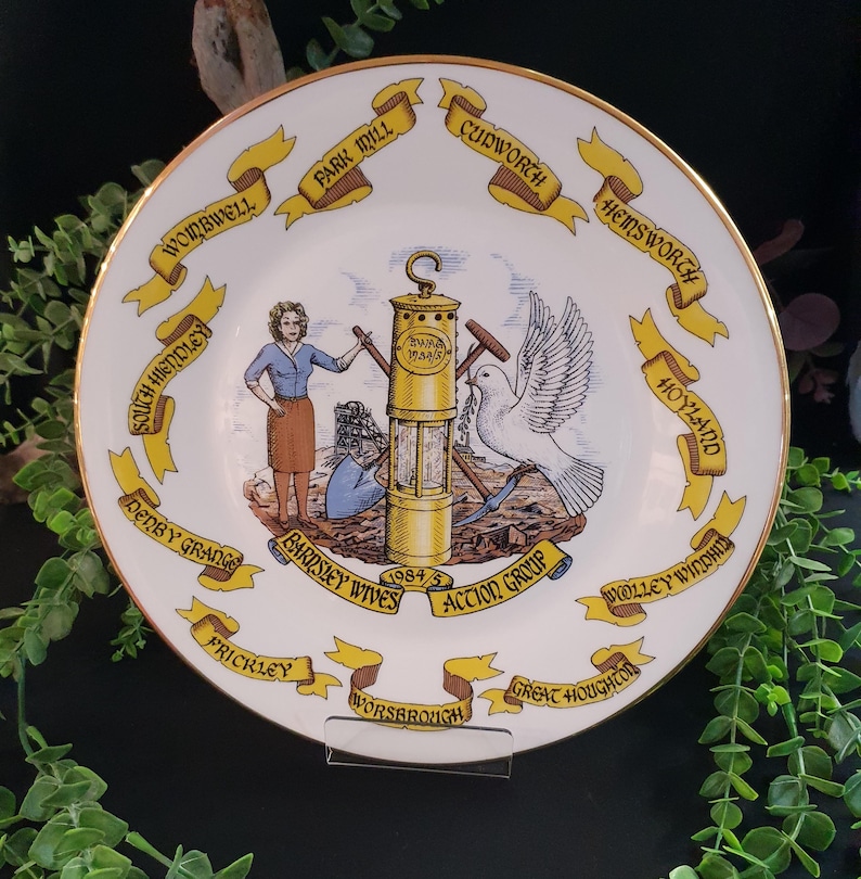 May include: A decorative white plate with a gold rim, featuring a central illustration of a miner's lamp, a dove, and figures. Yellow banners with town names encircle the plate. The text "BARNSLEY WIVES ACTION GROUP 1984/5" is also visible.