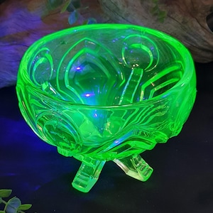 May include: A bright green glass bowl with a decorative pattern and three small feet. The glass has a textured surface and a neon-like appearance, possibly uranium glass. The bowl's design features curved and geometric shapes.