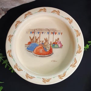 May include: Cream ceramic plate featuring a carnival scene with bunnies in bumper cars. The rim has a repeating pattern of running bunnies. The central illustration shows bunnies in bumper cars, flags, and the text "Barbara Vernon".