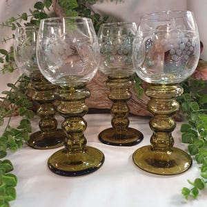 Op de afbeelding: Vier heldere wijnglazen met geëtste druivenontwerpen. De stelen zijn olijfgroen met een bolvormig ontwerp en de bases zijn rond. De glazen zijn gerangschikt op een wit oppervlak met groen gebladerte op de achtergrond.