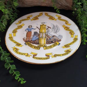 May include: A decorative white plate with a gold rim, featuring a central illustration of a miner's lamp, a dove, and figures. The plate is adorned with banners and text, including "BARDSLEY WIVES ACTION GROUP 1984/5".