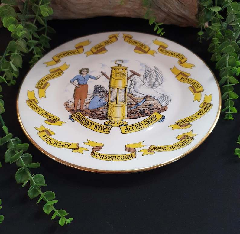 May include: A decorative white plate with a gold rim, featuring a central illustration of a miner's lamp, a dove, and figures. The plate is adorned with banners and text, including "BARDSLEY WIVES ACTION GROUP 1984/5".