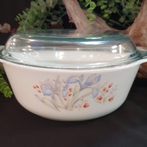 May include: A white casserole dish with a clear glass lid. The dish features a floral design with blue and orange flowers. The handles are white and the lid has a teal rim. A vintage kitchenware item.
