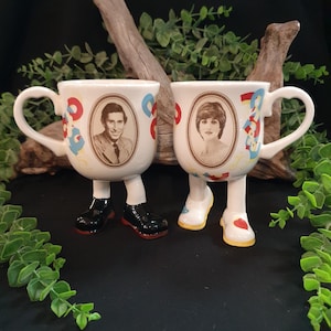 Puede incluir: Dos tazas de cerámica con retratos, uno de un hombre y otro de una mujer. Las tazas tienen cuerpos blancos con diseños florales y abstractos coloridos. Cada taza tiene un asa y está sostenida por piernas y pies, una con zapatos negros y la otra con zapatos blancos.