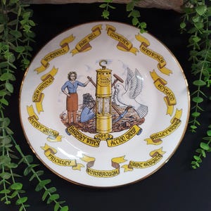 May include: A decorative white plate with a gold rim, featuring a central illustration of a mining lamp, a dove, and figures. The plate is encircled by yellow banners with various town names, including "Barnsley Wives Action Group 1984/5".
