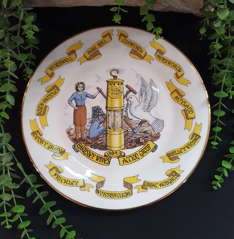 May include: A decorative white plate with a gold rim, featuring a central illustration of a mining lamp, a dove, and figures. The plate is encircled by yellow banners with various town names, including "Barnsley Wives Action Group 1984/5".
