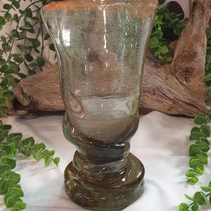 May include: A clear glass vase with a flared top and a decorative, swirling base. The rim of the vase has an orange tint. The base is a darker green colour. The vase is likely handmade and has a unique, organic shape.