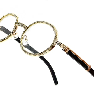 May include: Gold-rimmed eyeglasses with clear lenses. The round frames are encrusted with small, sparkling rhinestones. The temples are black with gold accents, and the glasses are set against a white background.