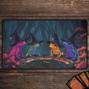 May include: A play mat featuring four colorful frogs wearing witch hats, playing cards in a forest setting. The frogs are pink, green, yellow, and blue. The mat has a dark background with the word "MAGIC" on a small box.