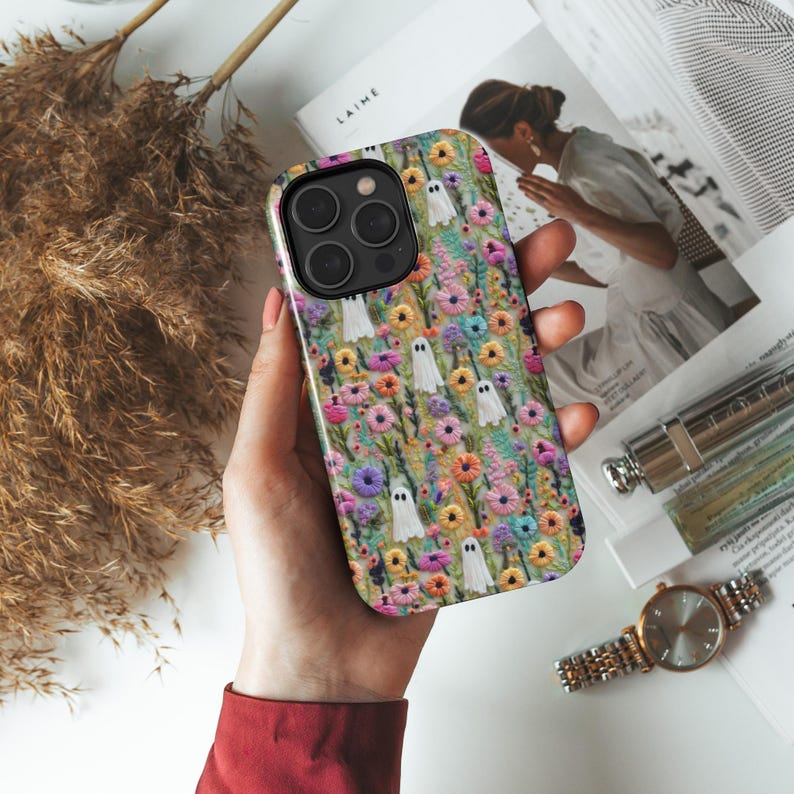 May include: A colorful phone case featuring a floral pattern with small white ghost illustrations. The case has a black camera module and is held in a person's hand. The background includes a magazine, watch, and dried plants.