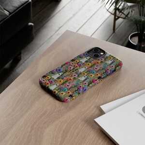 May include: A smartphone case featuring a colorful floral pattern with small white ghost figures. The case has a black camera lens cutout and is resting on a light-colored wooden surface.