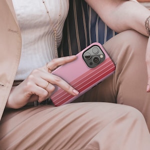 May include: A pink phone case with a gradient of red stripes. The case has a camera cutout and is held by a person wearing rings and a gold necklace. The case is a solid color with a series of horizontal stripes.