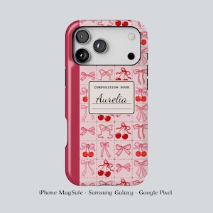 May include: A pink phone case with a pattern of bows and cherries. The case has a composition book design with the name "Aurelia" on it. The case is compatible with iPhone MagSafe, Samsung Galaxy, and Google Pixel phones.