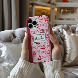 May include: A pink phone case with a pattern of bows and cherries. The case has a composition book design with the name "Aurelia" written on it. The case is held in a person's hands.