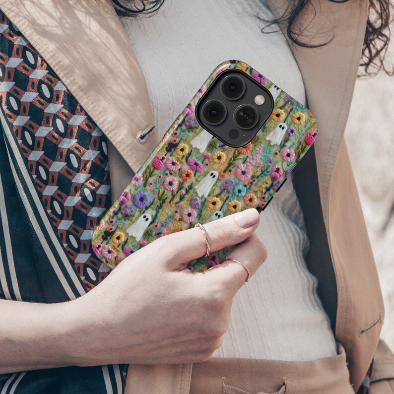 May include: A colorful phone case featuring an embroidered design of white ghosts and various flowers. The case has a black camera module and is held by a person. The flowers are in shades of pink, yellow, and blue.