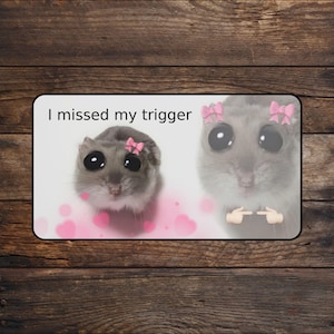 May include: A rectangular sign featuring two cartoon hamsters with large eyes and pink bows. The text "I missed my trigger" is at the top. Pink hearts are scattered around the hamsters, and one hamster is making a gesture with its paws.